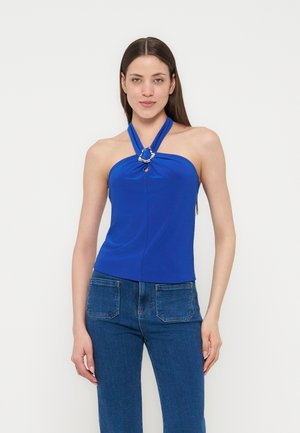 Γυναίκα που φοράει αμάνικο μπλε κοντό τοπ με λαιμόκοψη halter και ασημένια λεπτομέρεια δαχτυλιδιού, συνδυασμένο με ψηλόμεσο μπλε τζιν, στέκεται μπροστά σε απλό φόντο.