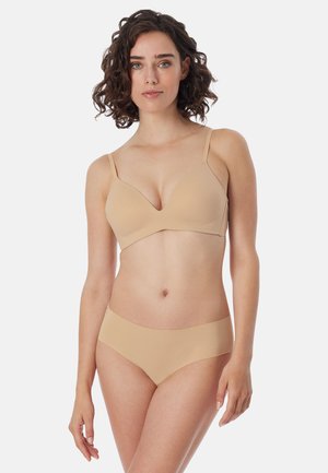 Kvinde med krøllet mørkt hår iført beige sømløs bh og matchende undertøj, vendt mod fronten mod ens hvid baggrund.