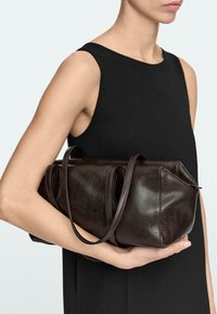 Femme en robe noire sans manches tenant un sac à main en cuir marron foncé avec deux bretelles, sur un fond clair uni.