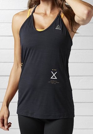 Frau trägt ein schwarzes Reebok-Top über einem gelben Sport-BH und steht vor einem weißen Holzwand-Hintergrund.