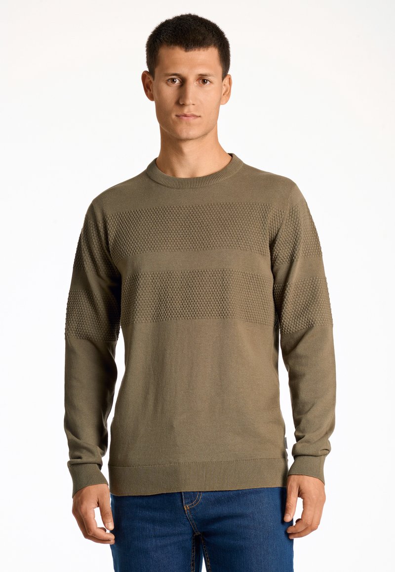 Maglione lavorato a maglia verde oliva con righe orizzontali testurizzate, scollatura rotonda e polsini a costine. Abbinato a jeans blu.