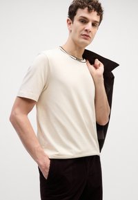T-shirt crème à manches courtes avec un col noir, assortie à un pantalon en velours côtelé marron foncé. La personne tient une veste.
