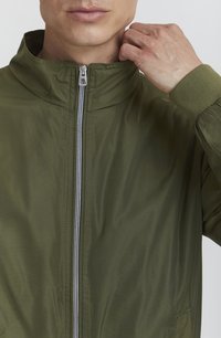 Olivgrüne, leichte Jacke mit hohem Kragen und silbernem Reißverschluss. Verfügt über elastische Bündchen und eine glatte, schimmernde Textur. Schlichtes, modernes Design.
