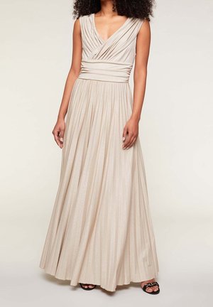 Femme portant une robe beige plissée sans manches avec une taille froncée et un col en V, associée à des sandales noires à bout ouvert.