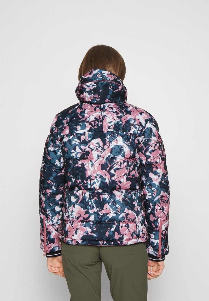 Personne portant une doudoune avec un motif floral rose, marine et blanc, debout de dos sur un fond uni.