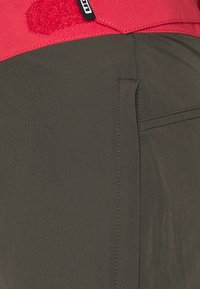 Czerwona tkanina o miękkiej teksturze łączy się z ciemnozieloną, gładką tkaniną. Wyróżniają się szczegóły szycia oraz pionowa kieszeń na zielonej części.