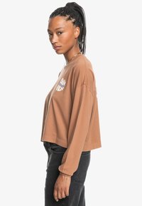 Bruine cropped sweatshirt met lange mouwen, voorzien van een ontwerp in witte en blauwe tekst, zachte textuur en een ontspannen pasvorm. Gecombineerd met zwarte broek.