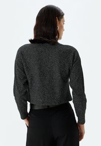 Gekürzter schwarzer Pullover aus einem strukturierten, glitzernden Stoff mit langen Ärmeln. Schwarze Hose mit einem taillierten Bund und zwei seitlichen Taschen.