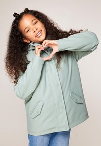 Giacca impermeabile verde menta con cappuccio, bottoni a pressione sul davanti e due tasche angolate. Il modello forma un cuore con le mani.
