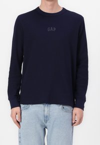 Marinblus långärmad ribbad tröja med rund hals och en subtil "GAP"-logotyp på bröstet, parat med ljusblå jeans.