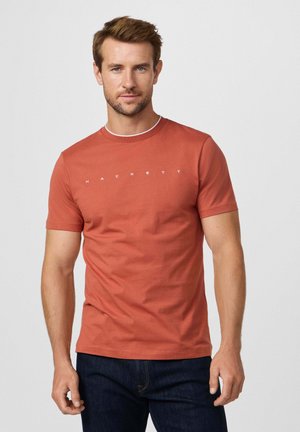 Camiseta de algodón roja con mangas cortas y cuello redondo, con texto impreso en blanco "HACKETT" en el frente, combinada con jeans de mezclilla oscura.