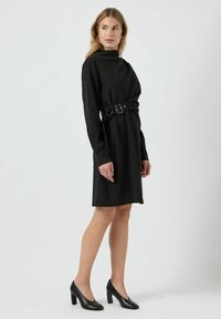 Černé šaty s dlouhým rukávem a vysokým výstřihem, stažené v pase opaskem, s elegantním siluetou a zkombinované s černými špičatými podpatky.