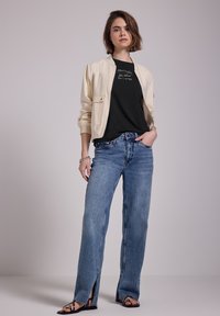 Cream bomber jacket, black graphic t-shirt, and light blue high-waisted jeans with side slits; model wearing black sandals, minimalist accessories.