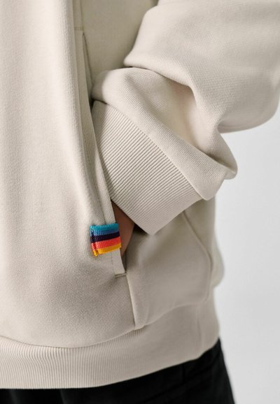 Manche de sweat-shirt beige avec poignet côtelé et main partiellement dans la poche, présentant une petite étiquette avec des rayures bleues, noires, rouges, orange et jaunes.
