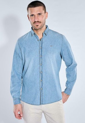 Man with short hair and beard wearing a buttoned light blue denim shirt and beige pants, standing with hand on hip against plain background.