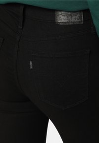 Jeans de denim negro con una vista en primer plano del bolsillo trasero, que presenta un patrón de costura distintivo y una etiqueta de tela con la marca.
