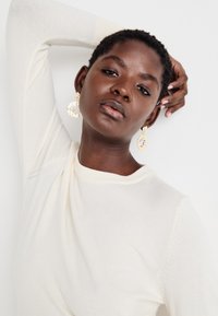 Pull en tricot blanc avec un col rond. Boucles d'oreilles pendantes au design circulaire avec des accents colorés et une finition texturée.