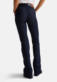 Tmavě modré denimové zvonové džíny s přilnavým pasem, kontrastním prošíváním a dvojitými zadními kapsami. Dokončeno hnědým koženým páskem.