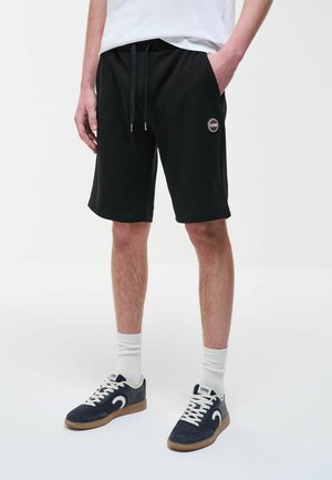 Persona che indossa pantaloncini neri con coulisse e patch con logo, calzini bianchi e sneakers blu navy con suole in gomma, in piedi contro uno sfondo chiaro uniforme.