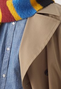 Beige trenchcoat med en skarp krage, lager över en ljusblå chambrayskjorta, med en flerfärgad randig ullsjal.