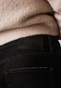 Jean en denim noir avec une ceinture texturée, dotée d'une pièce en cuir noir affichant le logo en relief "Calvin Klein Jeans".