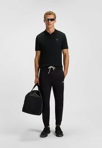 Polo noir avec logo, jogging noir avec cordon blanc, et baskets noires. Tenant un sac de sport noir ; des lunettes de soleil complètent le look.