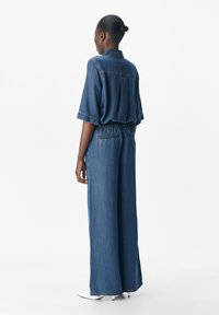 Femme de dos, portant une chemise en denim bleu et un jean ample avec des talons aiguilles blancs, contre un fond blanc uni.