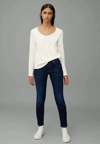Camisola branca de mangas longas com decote em V, combinada com jeans slim-fit azul-escuro e sapatilhas brancas. Tecido suave e design clássico. Sem padrões visíveis.