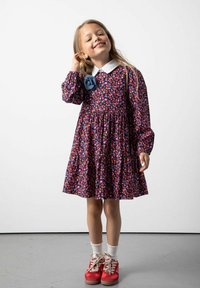 Kwiatowa sukienka w ciemnym niebieskim kolorze z białym kołnierzykiem, długimi rękawami i marszczonym stanikiem. Noszona z czerwonymi butami i białymi skarpetkami do kostek.