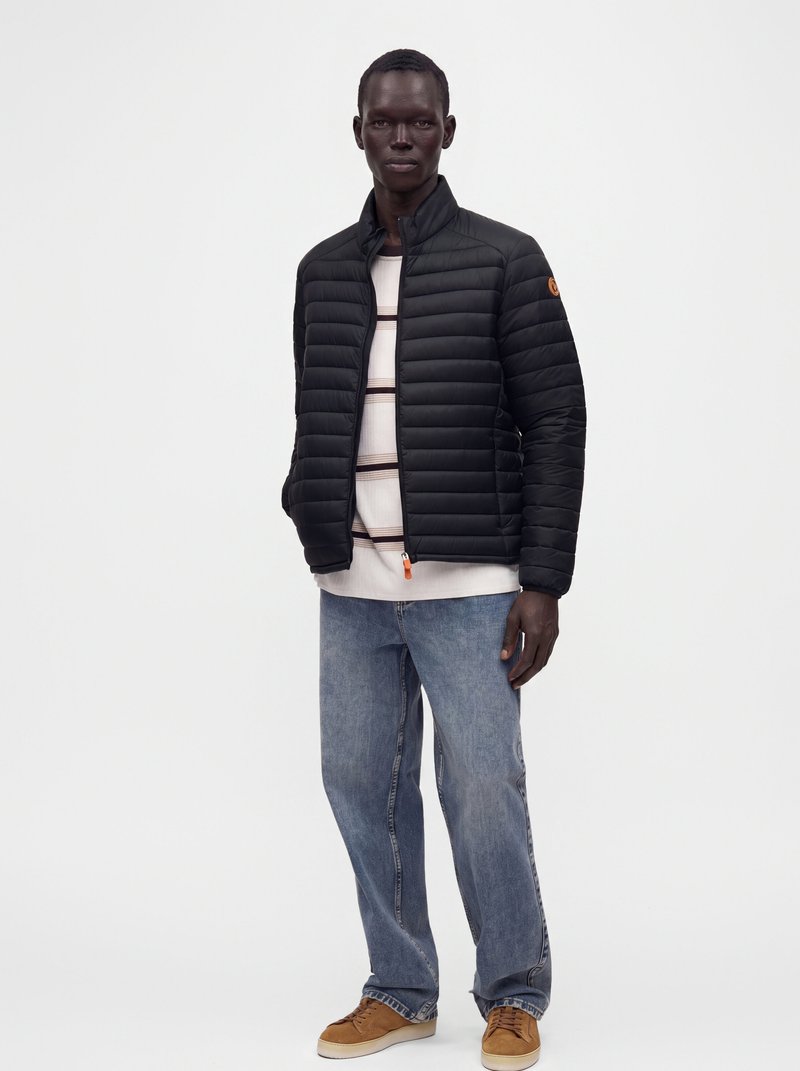 Homme portant une veste matelassée noire, une chemise rayée, un jean bleu et des baskets marron, debout sur fond blanc.