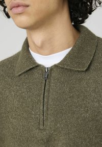 Pull en maille vert olive avec une fermeture éclair quart, un col et une finition texturée. Présente un t-shirt blanc visible sous l'encolure.