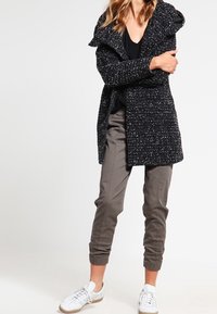 Cappotto lungo a pois bianco e nero con un ampio colletto, indossato sopra un top nero, abbinato a pantaloni oliva e sneaker bianche.