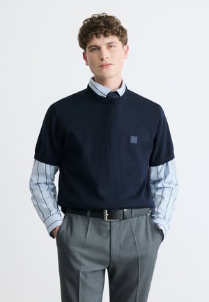 Joven con un suéter de manga corta azul marino sobre una camisa de rayas abotonada, pantalones grises y cinturón negro, posando con las manos en los bolsillos.