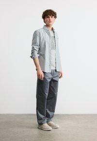 Νεαρός άνδρας με σγουρά μαλλιά, ντυμένος με ανοιχτόμπλε πουκάμισο με κουμπιά, λευκό κοντομάνικο με στάμπα, γκρι παντελόνι και λευκά αθλητικά παπούτσια, στέκεται σε γκρι δάπεδο.