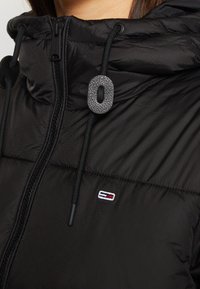 Doudoune noire matelassée avec capuche, dotée d'une languette texturée et d'un petit écusson logo avec des accents rouge, blanc et bleu.