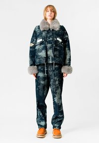 Chaqueta azul con estampado y cuello de piel gris, combinada con pantalones a juego y zapatillas naranjas. Presenta dos bolsillos frontales y cordón ajustable.