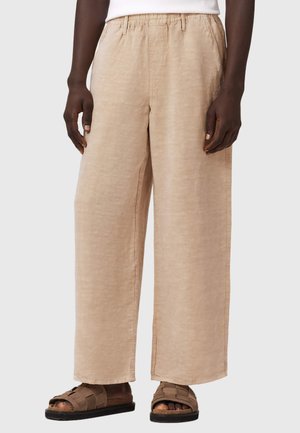 Person, die beige weit geschnittene Hosen mit elastischem Bund und braunen Sandalen mit offenen Zehen trägt, steht vor hellem Hintergrund.