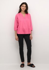 Blouse en lin rose à manches trois-quarts, décolleté en V et détails de boutons, associée à un jean noir ajusté et des sandales noires à enfiler.