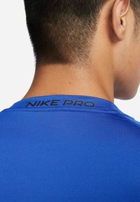 Nike Pro blå treningstrøye med rund hals og glatt tekstur. Merkenavnet er trykket i svart på baksiden av halsområdet.