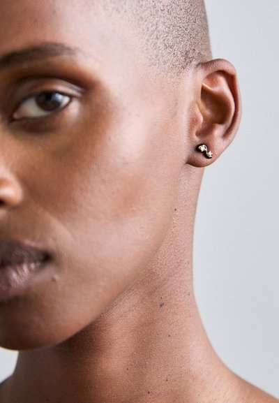 Gold-tone earrings in a curved design with small bead accents, worn in a close-up view; skin tone is deep brown against a neutral background.
