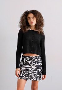 Μαύρο ριγέ κοντό κάρντιγκαν με μακριά μανίκια και κουμπιά μπροστά, συνδυασμένο με μια φούστα με ψηλή μέση σε ζέβρα, που διαθέτει υφασμάτινο μοτίβο.