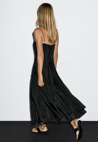 Sort maxikjole med spaghetti-stropper, subtil glans, flydende silhuet og samlet stofdetaljer, parret med flade sorte sandaler med stropper.