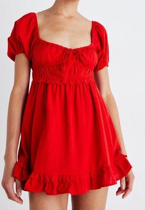 Kobieta w krótkiej czerwonej sukience z bufkami, marszczonym biustem i falbanami przy dolnej krawędzi na jednolitym białym tle.