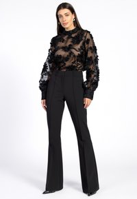 Vrouw in een doorschijnende zwarte blouse met bloemenborduursels en pofmouwen, zwarte hoge taille uitlopende broeken en zwarte puntige schoenen.