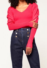 Donna con un top a maniche lunghe di un luminoso rosa fucsia con spalle a palloncino e jeans scuri a vita alta con bottoni decorativi, braccia incrociate in vita.