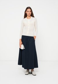 Cream ribbed cardigan with button front, paired with a dark navy pleated maxi skirt. The outfit is complemented with gray slip-on shoes.