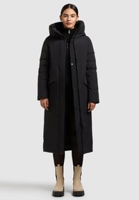 Sort pufferjakke med hætte, lang længde, frontlynlås, trykknapper og sidelommer; båret over en mørk trøje og nederdel.