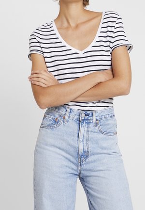 Henkilö, jolla on ristissä käsivarret, pukeutunut valkoiseen ja mustaraitaiseen V-kaulaiseen T-paitaan sekä korkeavyötäröisiin vaaleansinisiin farkkuihin yksinkertaista taustaa vasten.