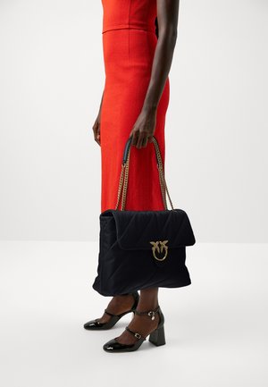 Borsa a tracolla nera trapuntata con hardware dorato e catene, caratterizzata da un design con pattina piegata. Indossata con un abito rosso aderente e tacchi neri.