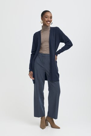 Femme souriante portant des boucles d'oreilles créoles en or, un col roulé marron, un cardigan bleu marine, un pantalon large bleu et des bottines à talons beiges, debout avec une main sur la hanche.